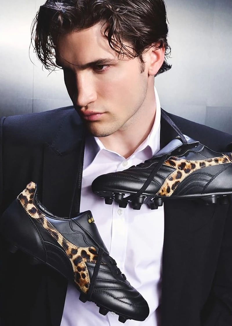 Max Knott with wet-look brown hair in a black tailored blazer and white dress shirt, holding up the Dolce & Gabbana x Diadora Brasil in black leather with leopard pony hair side panels