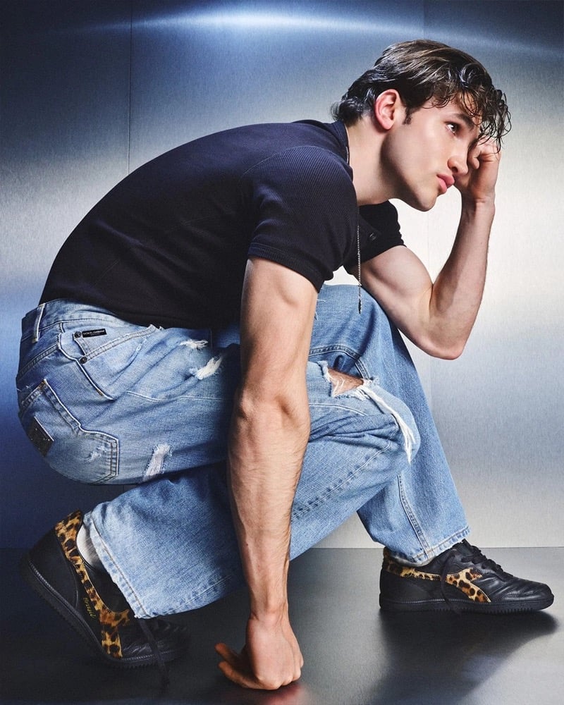 Max Knott crouched in a black ribbed Dolce & Gabbana t-shirt and shredded light wash blue jeans with a Dolce & Gabbana waistband label, wearing the leopard pony hair Brasil sneaker against a brushed silver backdrop