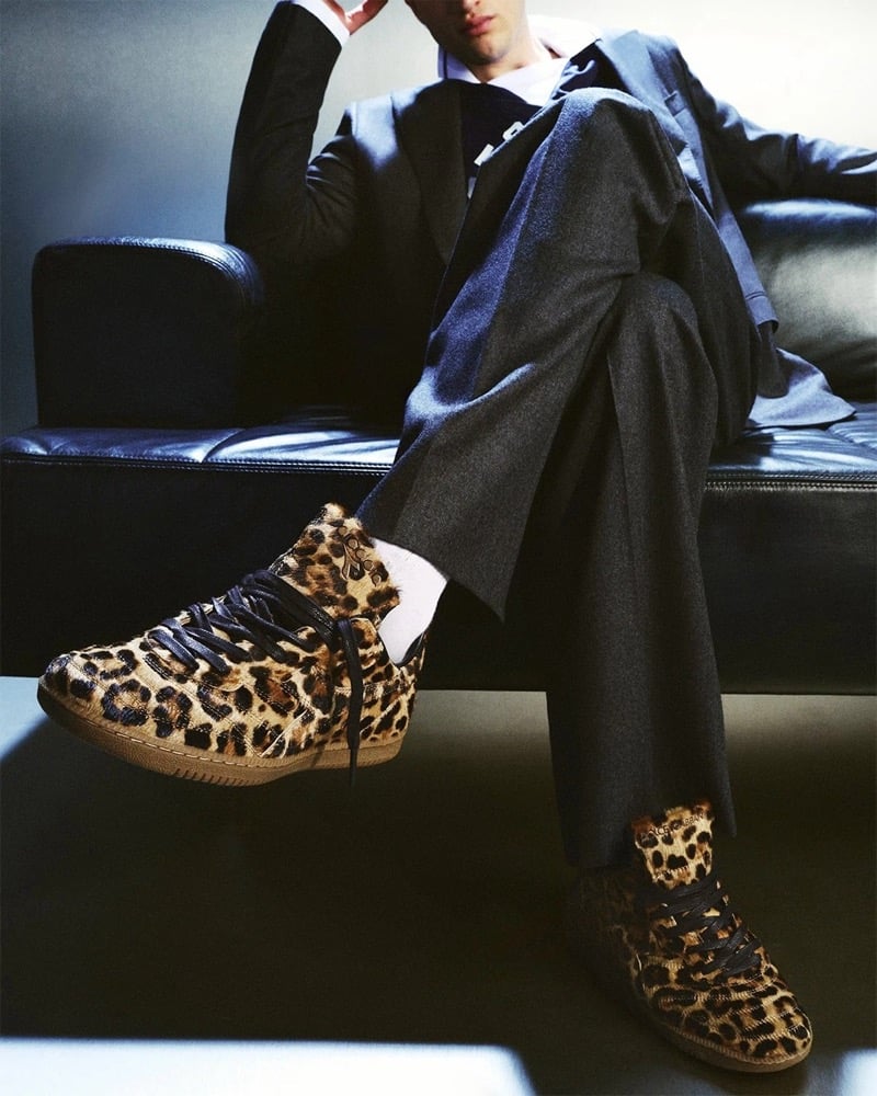 Max Knott reclined on a black leather sofa in a charcoal gray wool suit and dress shirt with a navy knit tie, wearing leopard pony hair Dolce & Gabbana x Diadora Brasil high-top sneakers with white sport socks branded in gold lettering