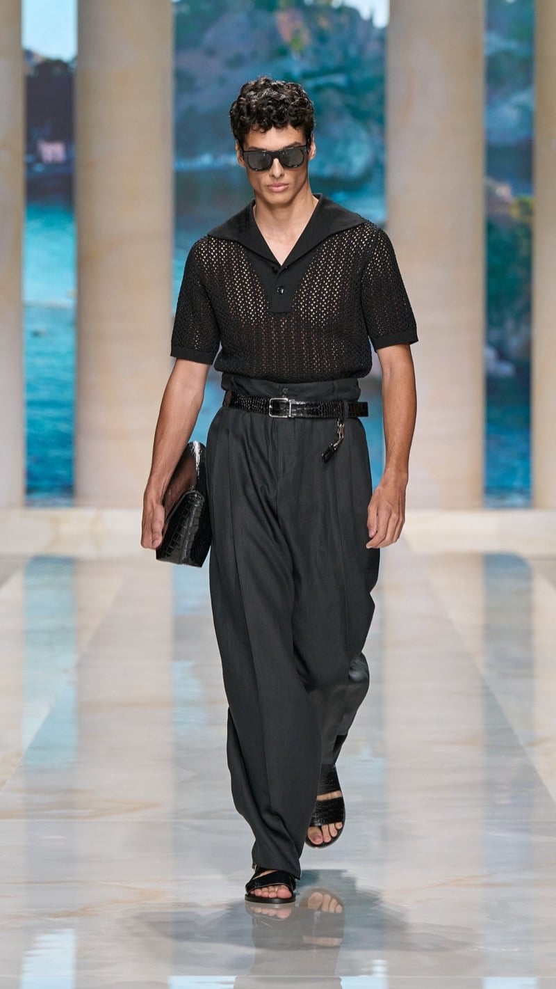 Model in dark sunglasses wears a dark openwork mesh sailor-collar polo with wide, high-waisted pleated dark trousers, a braided belt with key fob, dark sandals, and carries a dark croc-embossed leather clutch