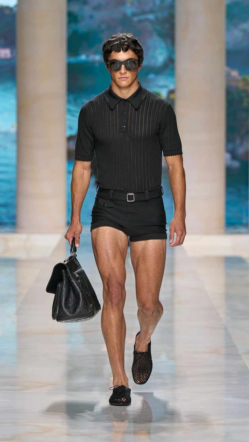 Model in shield sunglasses wears a ribbed dark short-sleeve polo tucked into slim belted dark shorts, paired with crochet mesh derby shoes and a slouchy dark leather top-handle bag