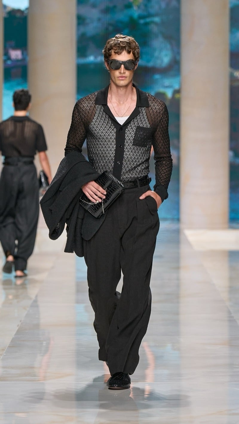 Model in aviator sunglasses wears a sheer dark diamond-mesh camp-collar shirt over a tank, paired with wide pleated dark linen trousers and dark crochet loafers, carrying a folded jacket and a dark croc-embossed clutch