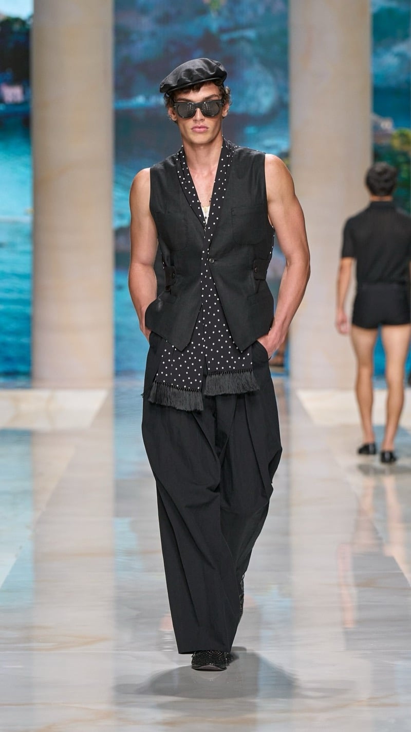 Model in a leather flat cap and sunglasses wears a sleeveless dark waistcoat over a long fringed polka dot scarf, paired with very wide pleated dark linen trousers