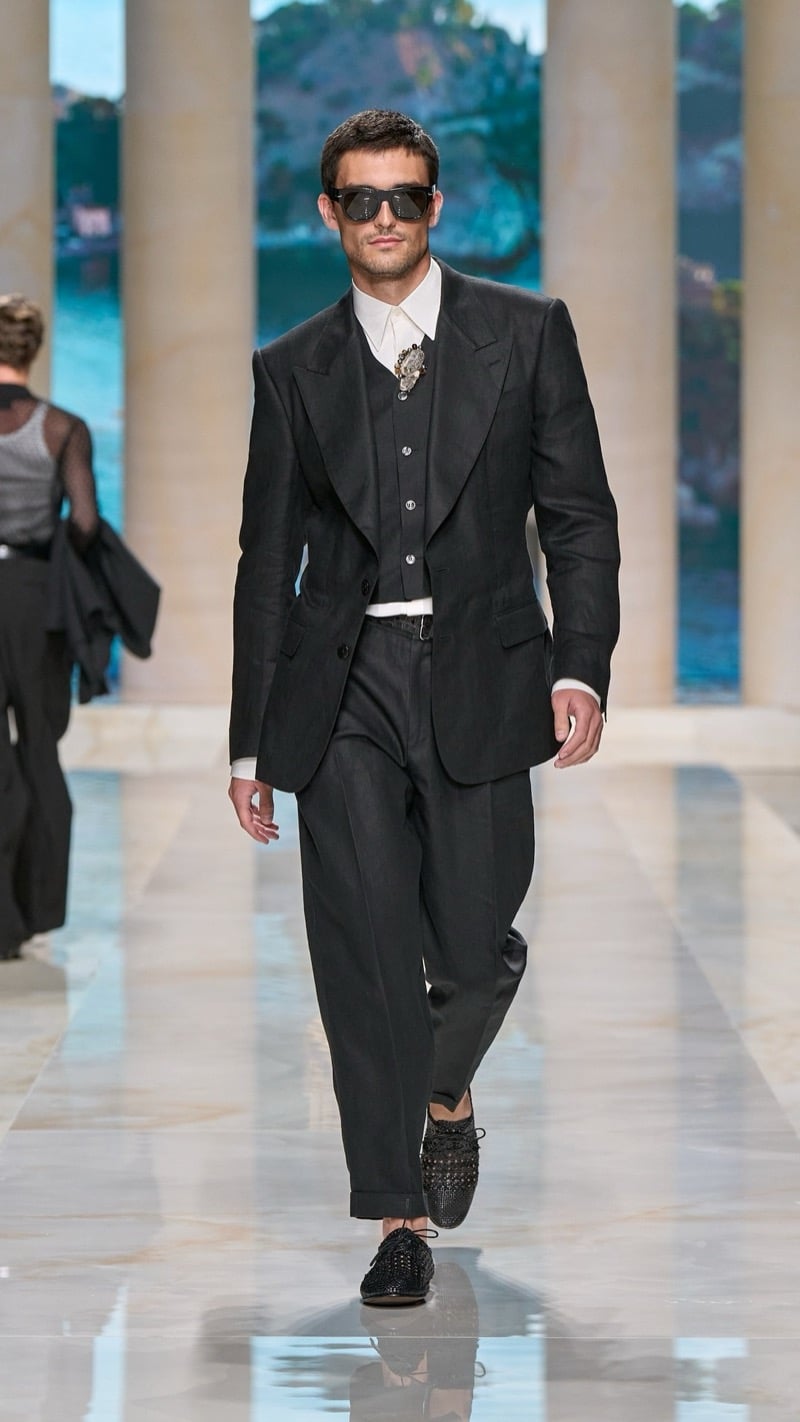 Model in aviator sunglasses wears a peak-lapel dark linen suit with matching waistcoat, light dress shirt, a jeweled brooch at the collar, and lace-up woven dark dress shoes
