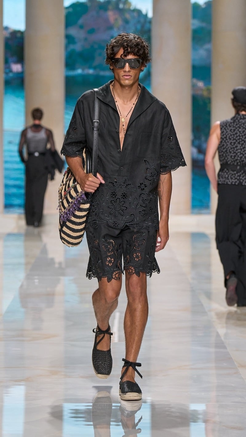 Model in sunglasses wears a dark scalloped cutwork-eyelet camp shirt and matching shorts set, paired with dark ankle-tie espadrilles and a striped raffia tote with a leather strap