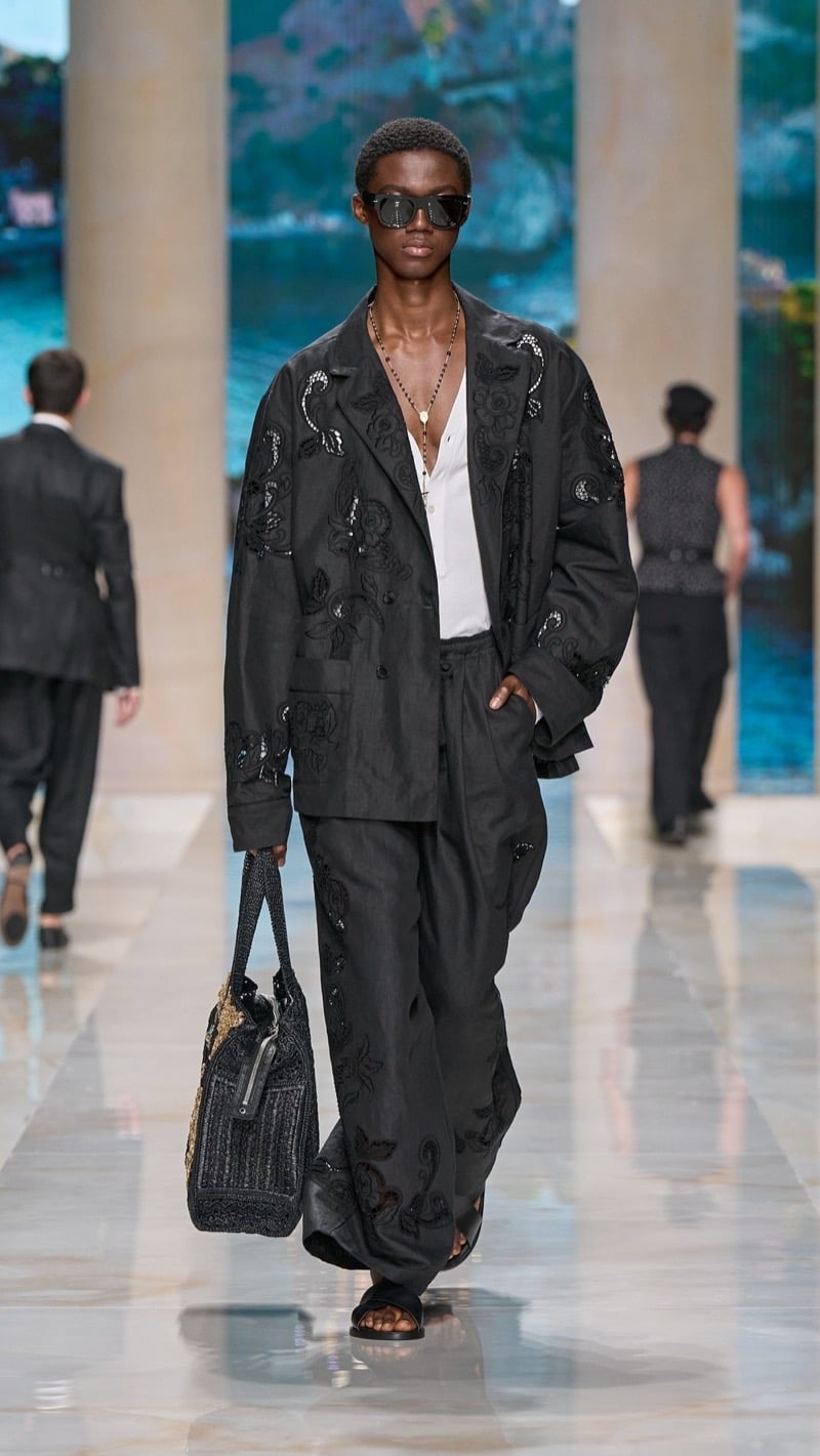 Model in flat-top sunglasses wears an oversized dark linen jacket and wide trousers covered in floral cutwork embroidery, layered over a draped light blouse, with dark slide sandals and a crochet tote