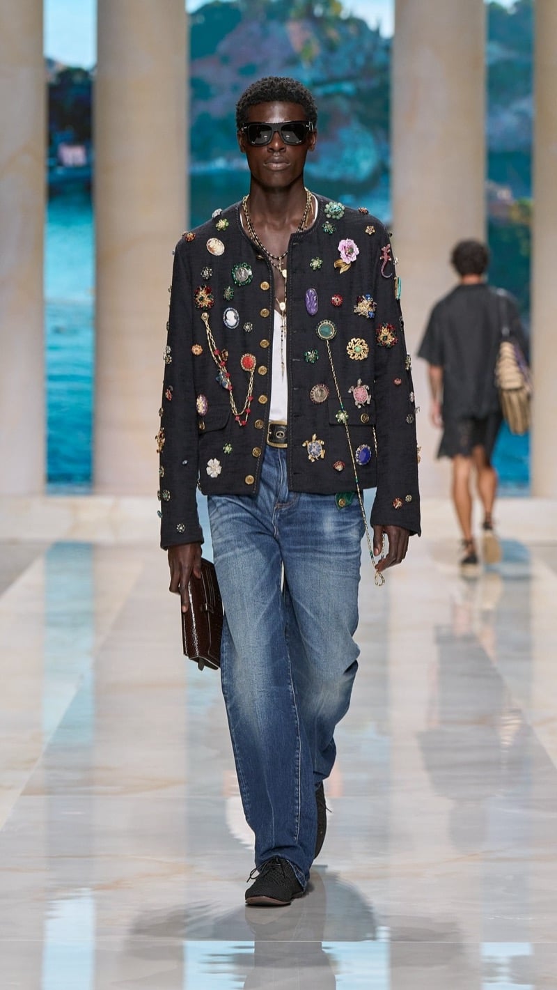 Model in sunglasses wears a dark cropped jacket densely covered in colorful jeweled brooches, cameos, and charm chains, layered over a tank, paired with light wash baggy jeans and dark woven derbies