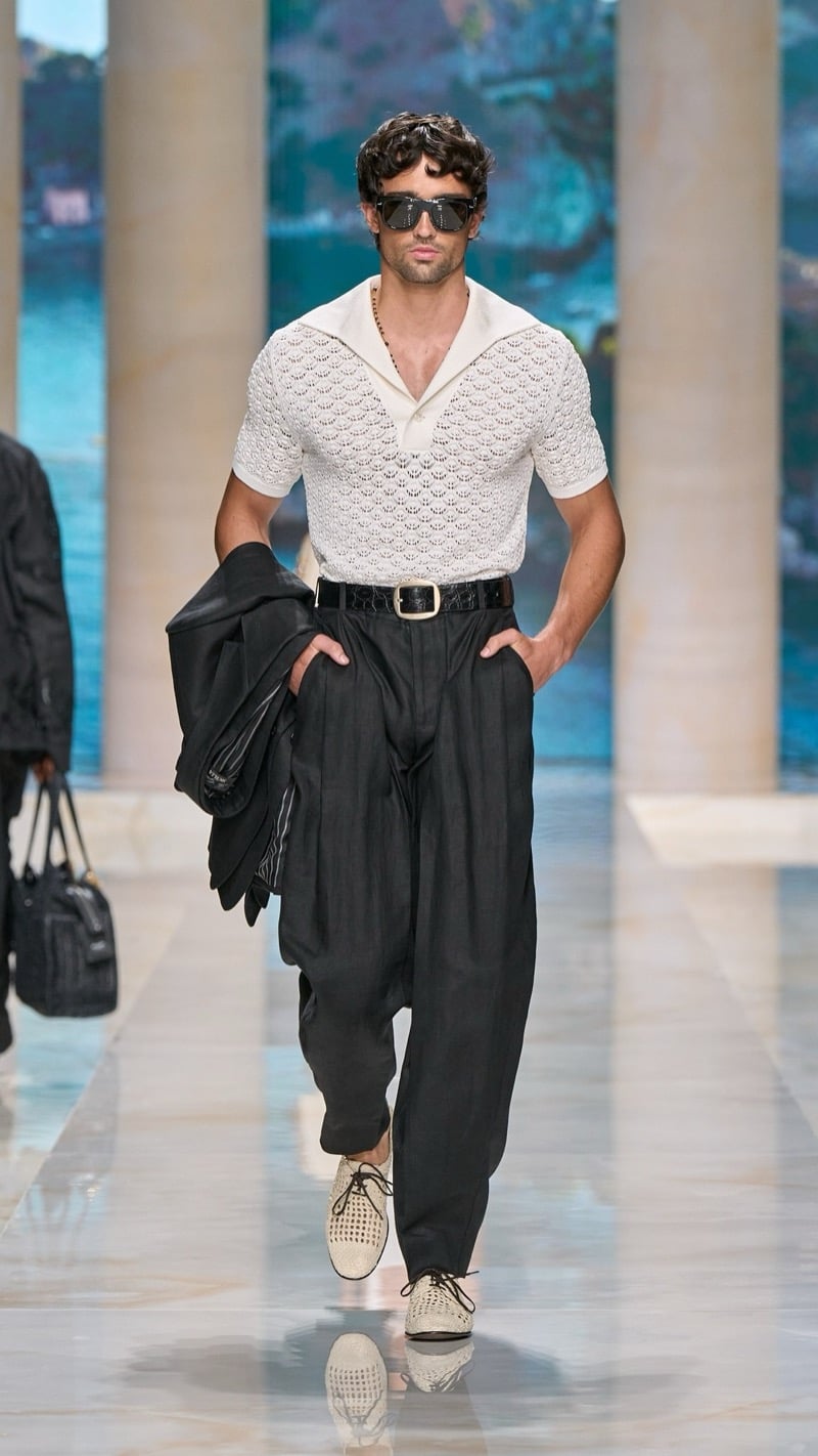 Model in flat-top sunglasses wears a short-sleeve openwork crochet polo with a sailor collar tucked into wide pleated dark linen trousers with a croc-embossed belt, paired with light woven mesh derbies