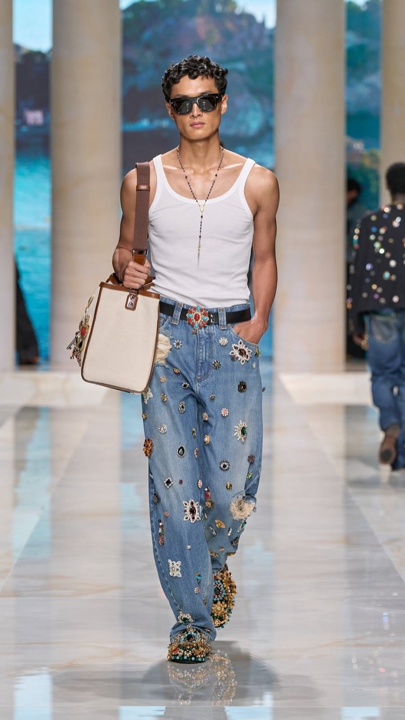 Model in sunglasses wears a ribbed light tank with a rosary necklace and light wash relaxed jeans densely jeweled with multicolored stones and brooches, paired with a heavily jeweled shoe and a canvas and leather tote