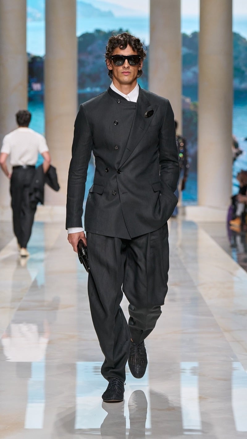 Model in square sunglasses wears a double-breasted band-collar dark linen suit with a peak-lapel construction, light dress shirt, jeweled brooch at the chest, and dark woven raffia derbies