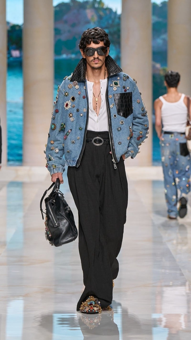 Model in shield sunglasses wears a denim bomber jacket densely covered in jeweled brooches with a quilted leather collar, layered over a light shirt, paired with wide pleated dark trousers, an ornate buckled belt, jeweled slippers, and a dark leather top-handle bag