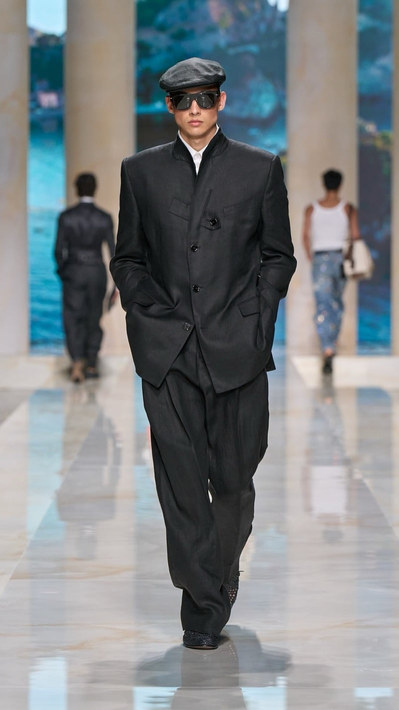 Model in a leather flat cap and shield sunglasses wears a band-collar dark linen overcoat-style jacket with a light dress shirt and wide pleated dark trousers, paired with dark woven derbies