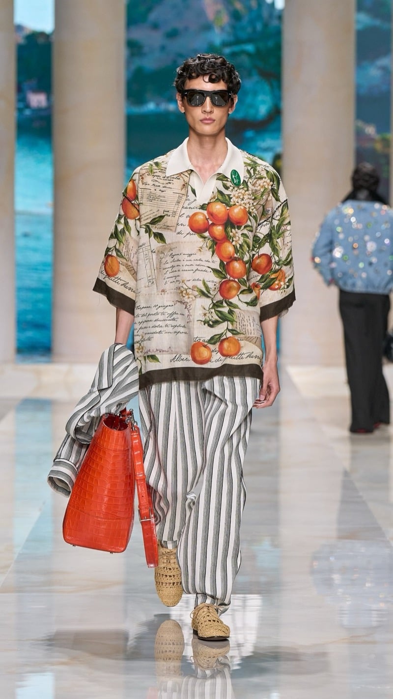 Model in sunglasses wears a short-sleeve camp shirt printed with a Sicilian citrus and postcard motif over wide pinstripe linen pajama pants, paired with raffia derbies and an orange croc-embossed tote with a striped robe