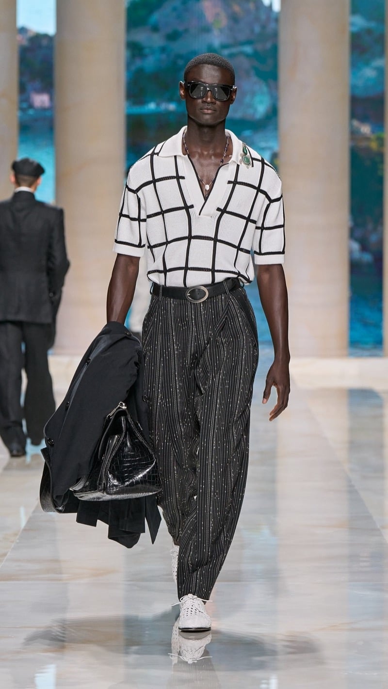 Model in sunglasses wears a light short-sleeve openwork knit polo with a dark windowpane pattern, tucked into wide pleated dark pinstripe trousers, paired with woven crochet sneakers and a folded jacket with a dark croc bag