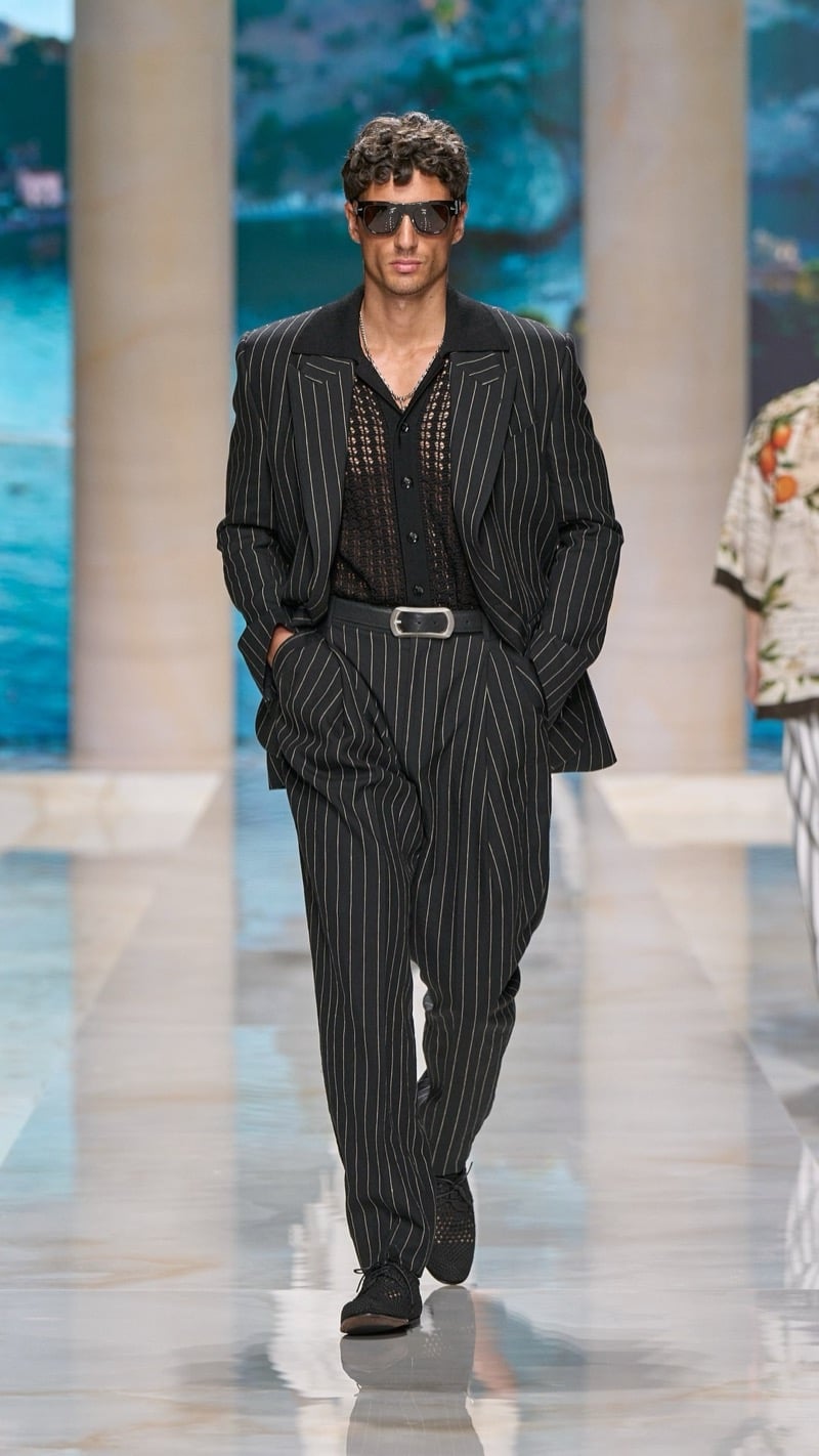 Model in shield sunglasses wears a peak-lapel pinstripe dark linen suit with a wide-set jacket, layered over a dark openwork knit shirt with a buckled belt, paired with dark woven derbies