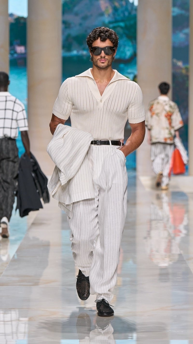 Model in shield sunglasses wears a ribbed short-sleeve light knit polo tucked into wide pleated light pinstripe trousers with a dark belt, paired with dark woven derbies and a folded pinstripe jacket
