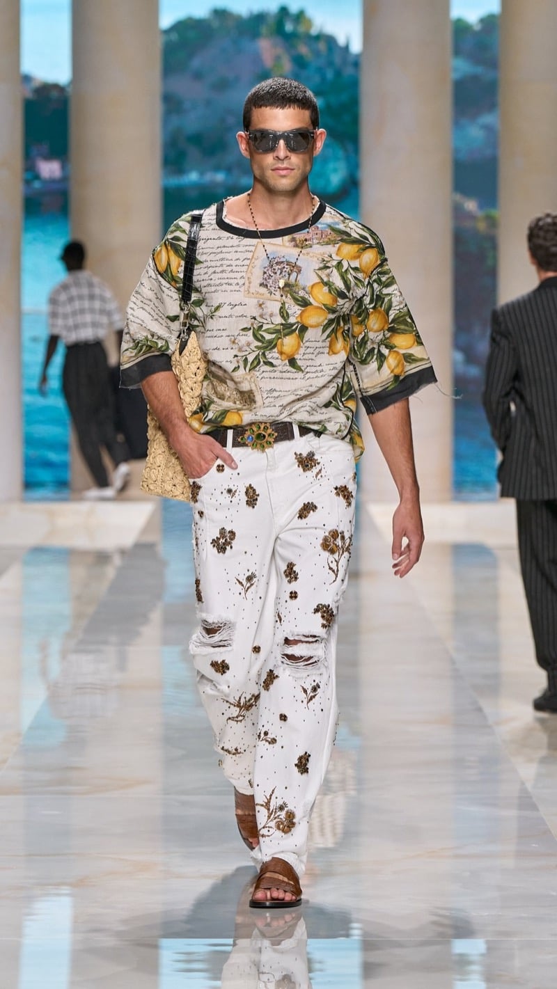 Model in sunglasses wears an oversized short-sleeve shirt printed with lemons and Sicilian script, paired with light distressed jeans densely embroidered with golden floral motifs, jeweled buckle belt, brown sandals, and a crochet tote
