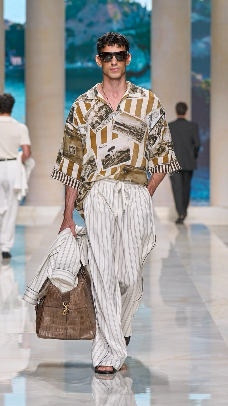 Model in shield sunglasses wears a short-sleeve camp shirt printed with vintage postcard imagery in tan and ivory, paired with wide drawstring pinstripe linen trousers, dark thong sandals, and a tan croc-embossed travel bag