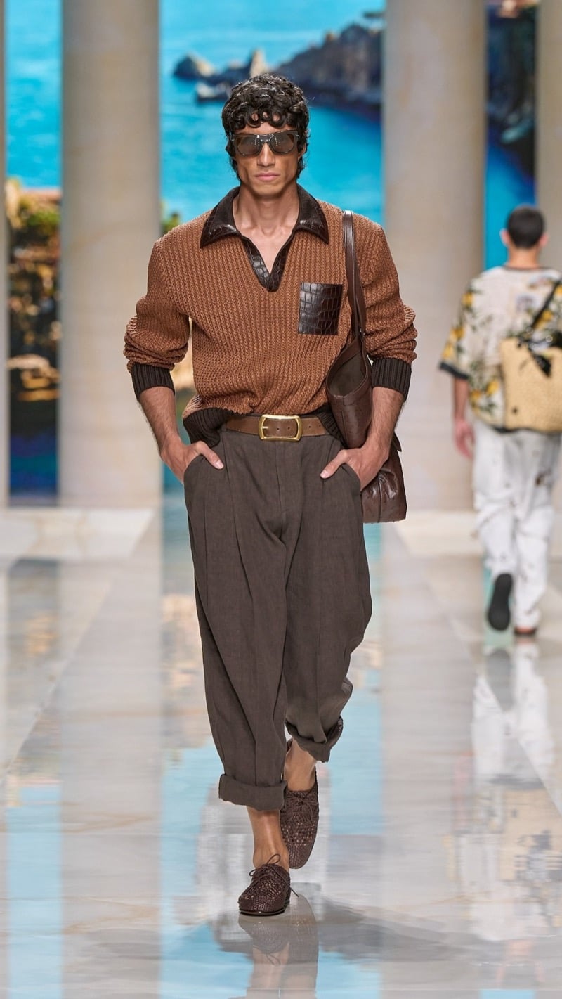 Model in shield aviator sunglasses wears a tobacco openwork knit polo with a dark croc-embossed collar and pocket trim, paired with wide pleated taupe linen trousers, a brown leather belt, dark woven derbies, and a brown leather bag