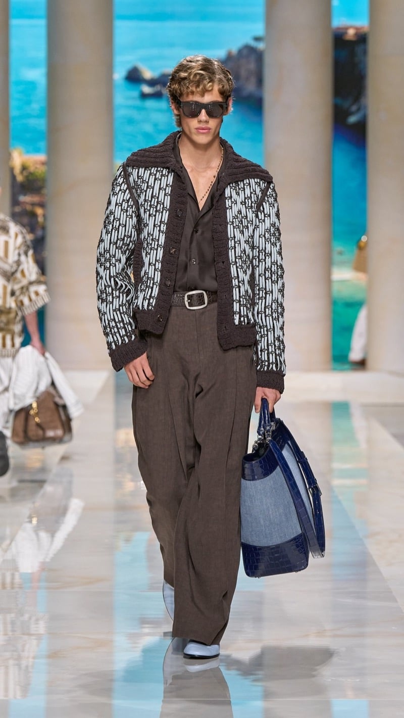 Model in shield sunglasses wears a chunky knit-trimmed monogram-print cardigan over a dark linen shirt, paired with wide pleated taupe linen trousers and a deep blue croc-embossed top-handle travel bag