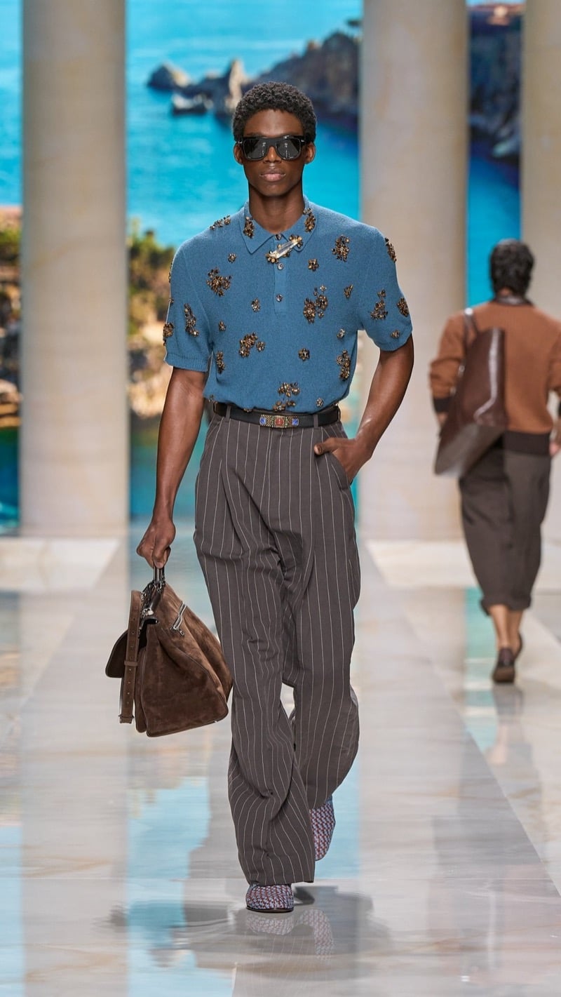 Model in shield sunglasses wears a slate blue short-sleeve knit polo scattered with jeweled appliques, tucked into wide pleated taupe pinstripe trousers, paired with a jeweled buckled belt, embroidered shoes, and a brown suede top-handle bag