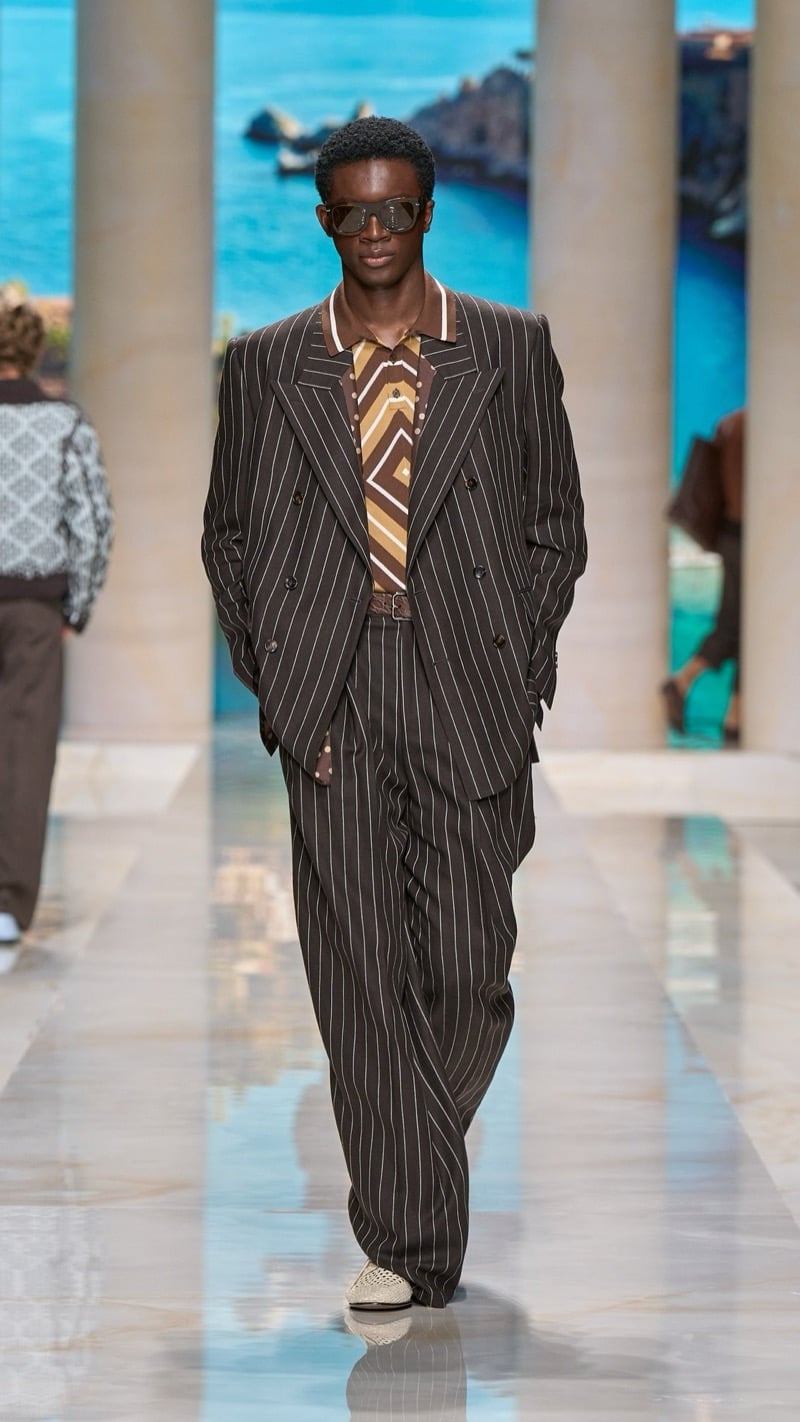 Model in flat-top sunglasses wears an oversized double-breasted dark brown pinstripe suit with peaked lapels, layered over a graphic-print knit polo, paired with crochet raffia derbies