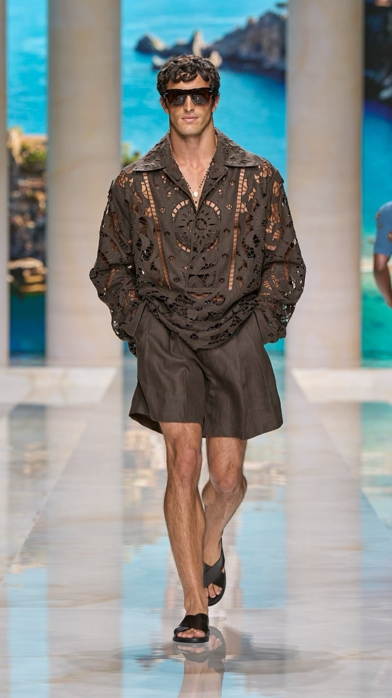 Model in shield sunglasses wears an oversized brown linen cutwork-embroidered shirt with wide pleated brown linen shorts, paired with dark slide sandals