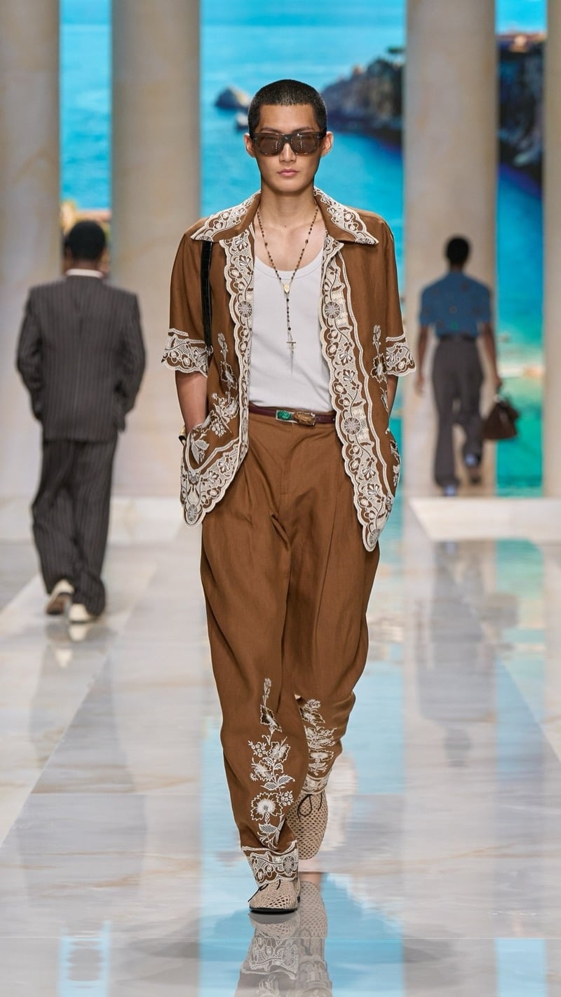 Model in sunglasses wears a tobacco camp-collar shirt with lace and embroidery trim, layered over a tank, paired with tobacco pleated trousers embroidered with floral motifs, an ornate belt buckle, and crochet raffia shoes
