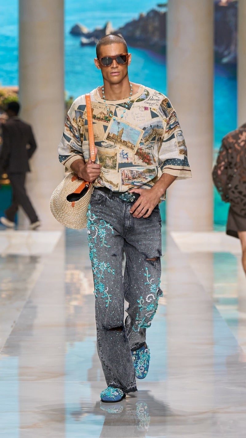 Model in tortoise sunglasses wears an oversized short-sleeve postcard-print tee over light wash distressed jeans densely embroidered with turquoise floral motifs, paired with jeweled turquoise shoes and a straw bag with an orange strap