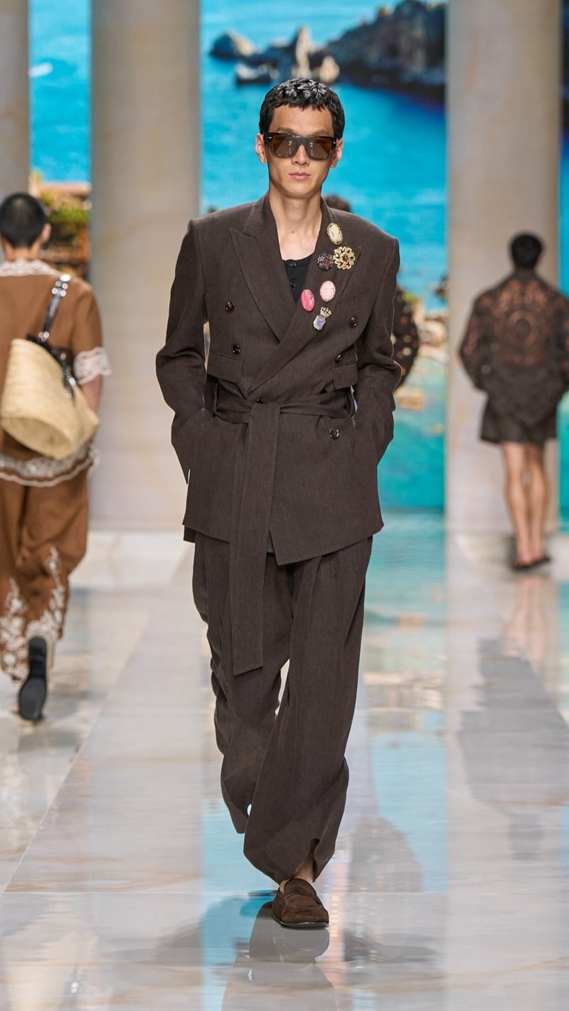 Model in shield sunglasses wears a double-breasted belted dark brown linen suit with peak lapels and a tied waist, layered over a dark tank decorated with multicolored cameo and gem brooches, paired with brown suede loafers