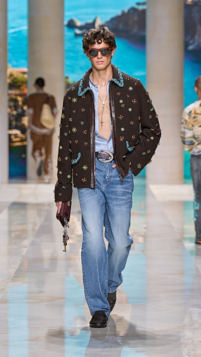 Model in sunglasses wears a brown jacket densely embellished with jewel and floral motifs and chenille embroidery, layered over an open shirt, paired with an ornate belt buckle, faded blue jeans, and brown loafers