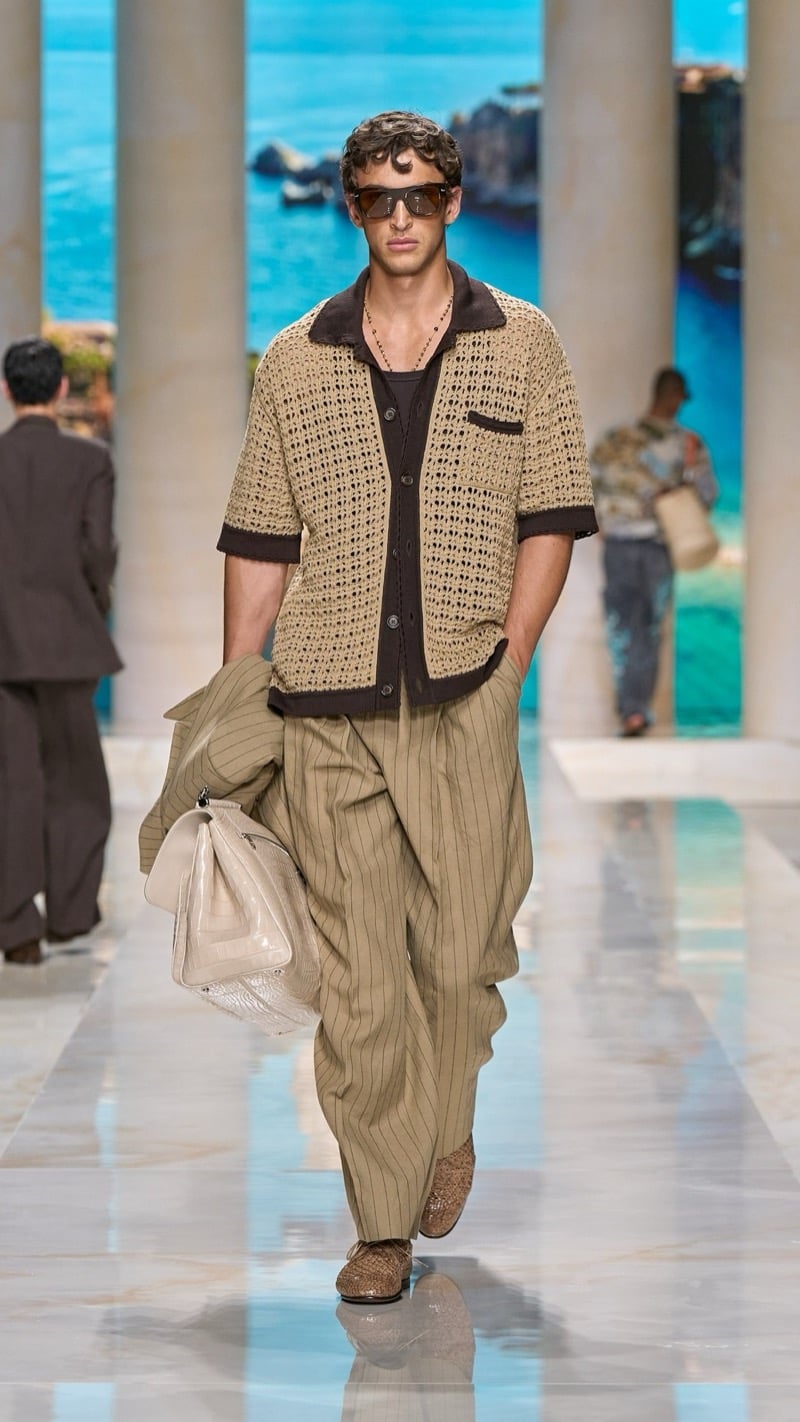 Model in sunglasses wears a short-sleeve openwork raffia knit cardigan with dark trim, paired with wide pleated tan pinstripe trousers and crochet raffia loafers, carrying a folded pinstripe jacket and a pale croc-embossed travel bag