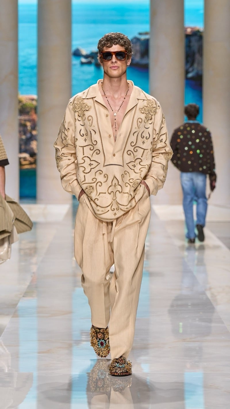 Model in tortoise sunglasses wears a sand camp-collar linen shirt and matching wide trousers densely embroidered with metallic baroque scrollwork, paired with elaborately jeweled brocade slippers