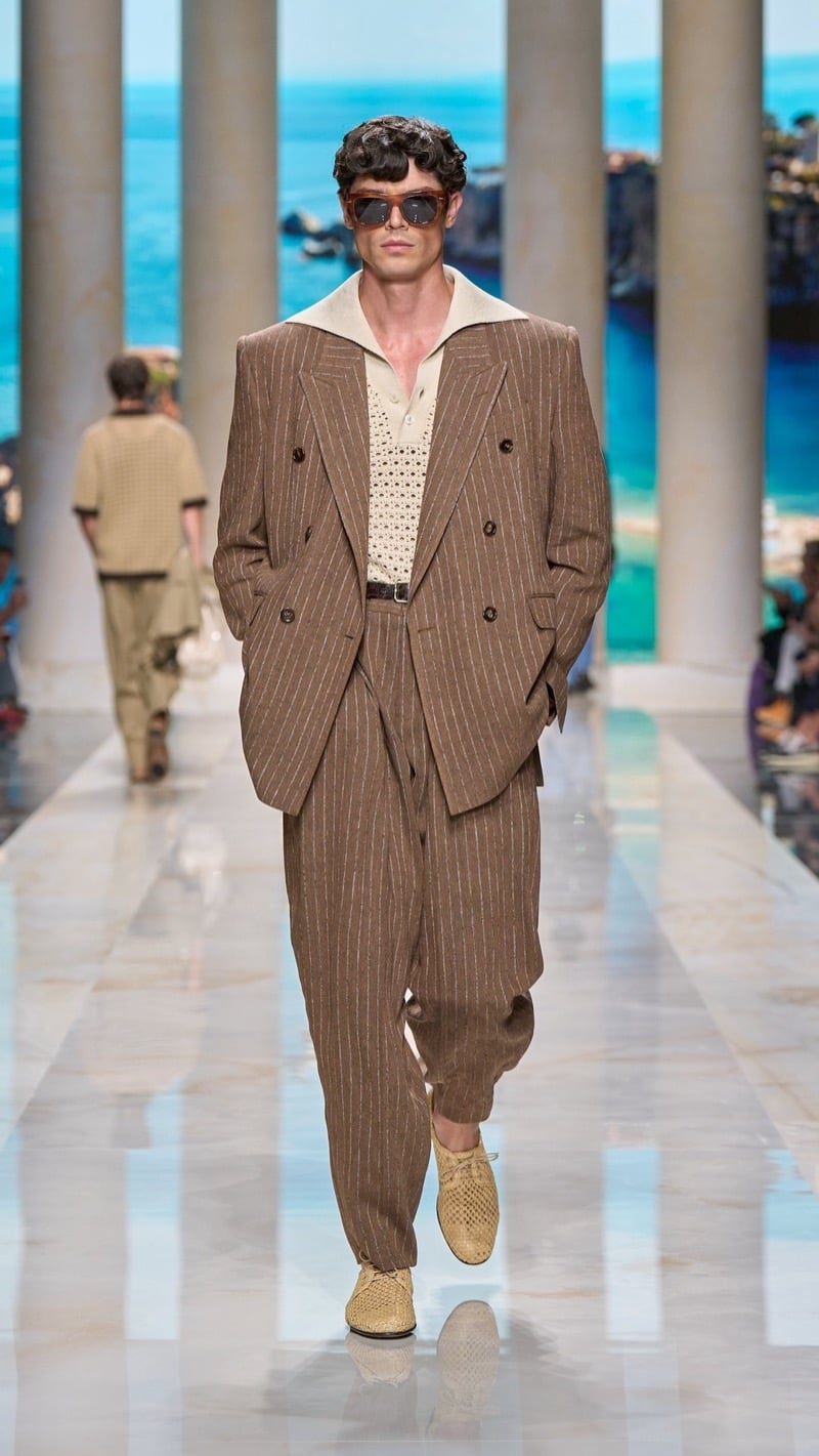 Model in tortoise sunglasses wears an oversized double-breasted brown pinstripe linen suit with peaked lapels, layered over an openwork raffia knit polo with a sailor collar, paired with raffia derbies