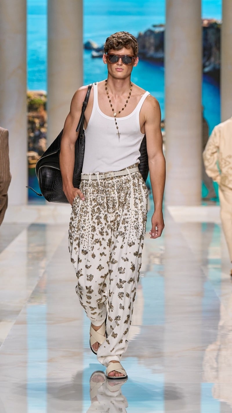 Model in shield sunglasses wears a ribbed light tank with a rosary necklace, tucked into wide pleated drawstring trousers densely beaded with metallic floral clusters, paired with crossed-strap sandals and a dark croc-embossed travel bag