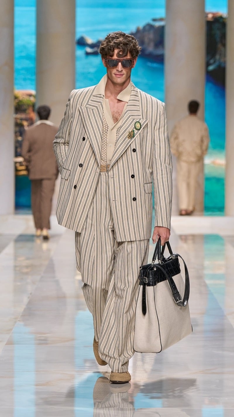 Model in sunglasses wears an oversized double-breasted striped linen suit with peaked lapels, layered over an openwork knit with jeweled brooch, paired with raffia derbies and a structured tote with dark croc trim