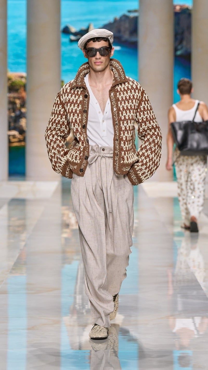 Model in a flat cap and aviator sunglasses wears a brown and ivory chunky houndstooth knit bomber over a light henley, paired with wide pleated drawstring oatmeal linen trousers and crochet raffia slip-on shoes
