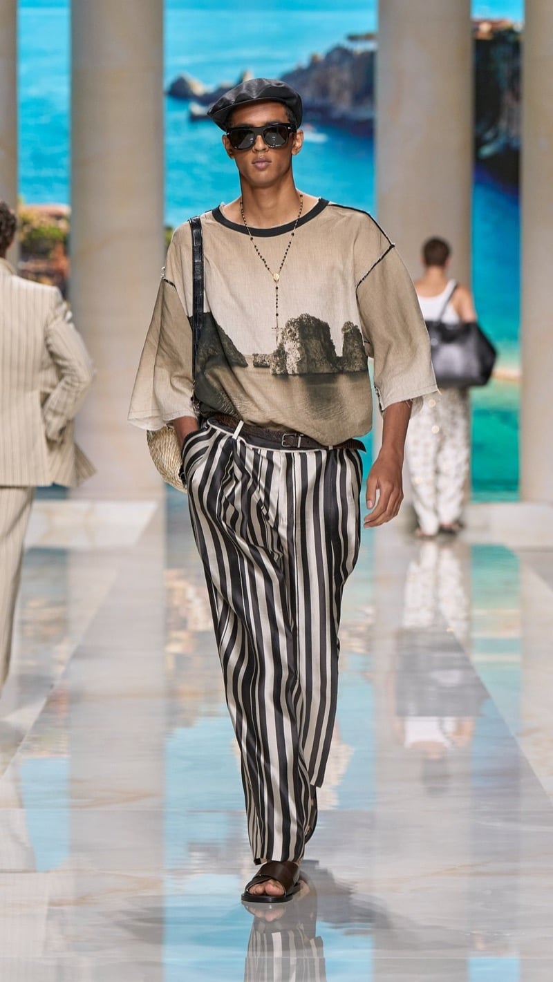 Model in a leather flat cap and sunglasses wears a relaxed light linen tee printed with a Sicilian sea-stack landscape, paired with wide pleated dark and ivory awning-striped silk trousers, a dark belt, dark thong sandals, and a raffia bag