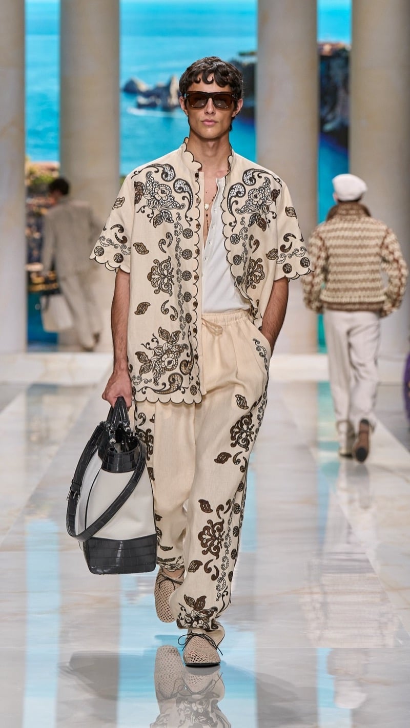 Model in shield sunglasses wears a stand-collar short-sleeve linen shirt and matching wide drawstring trousers densely printed with brown baroque embroidery motifs, paired with light raffia derbies and a dark croc-embossed bucket bag