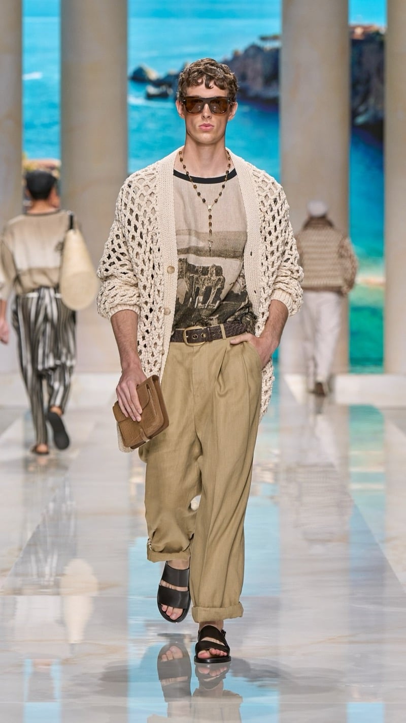 Model in flat-top sunglasses wears a relaxed graphic print tee under an oversized openwork raffia cardigan, paired with wide pleated khaki linen trousers, a wide leather belt, and dark slide sandals, holding a small suede pouch