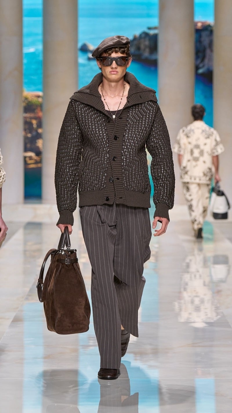 Model in a leather flat cap and sunglasses wears a chunky knit cargo cardigan in deep taupe, paired with wide pleated pinstripe trousers, dark loafers, and a brown suede top-handle bag