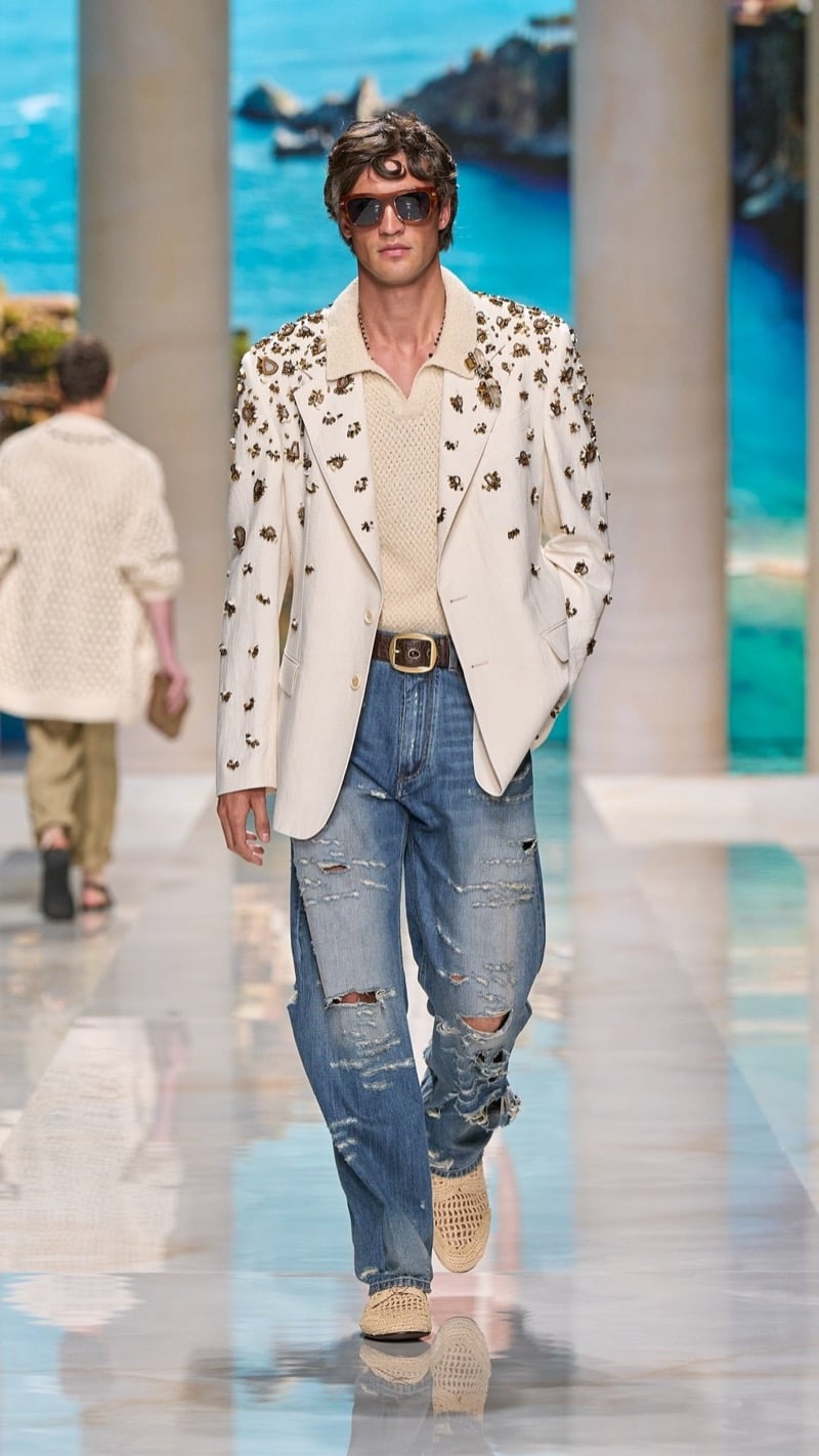Model in tortoise sunglasses wears an ivory blazer densely embellished with metallic gold floral appliques over an openwork knit polo, paired with an ornate buckled belt and distressed light wash jeans with crochet shoes