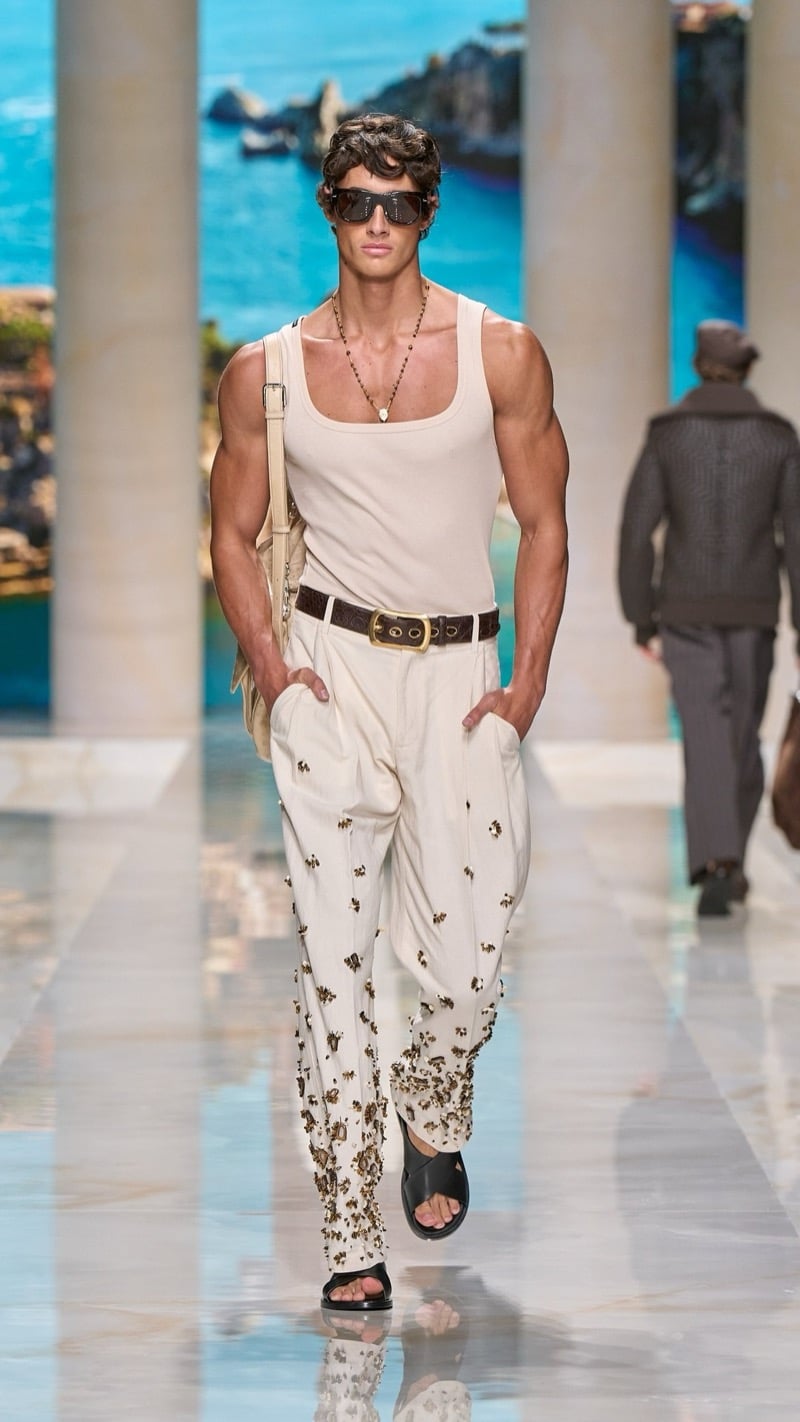 Model in shield sunglasses wears a ribbed ivory tank with a long bead chain, tucked into wide pleated ivory linen trousers densely embellished with bronze floral clusters, paired with a wide buckled belt, dark slide sandals, and a tan tote