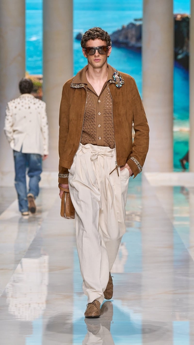 Model in tortoise aviator sunglasses wears a tobacco suede zip jacket with jeweled collar and cuff trim over a brown openwork knit cardigan, paired with wide pleated ivory linen drawstring trousers and brown crochet derbies