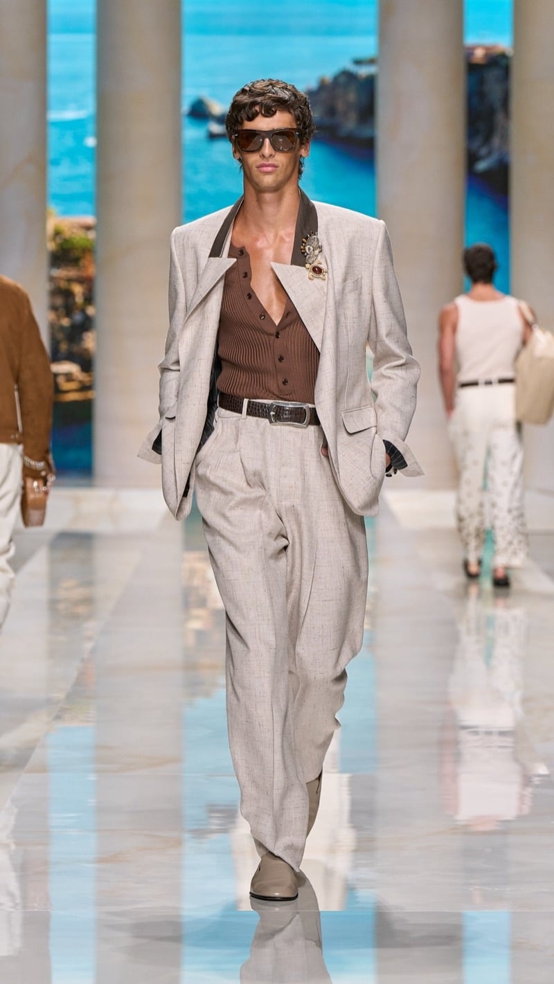 Model in tortoise sunglasses wears an oatmeal linen blazer and a jeweled brooch over a ribbed tobacco knit cardigan, paired with wide pleated oatmeal linen trousers, a dark croc-embossed belt, and pale loafers