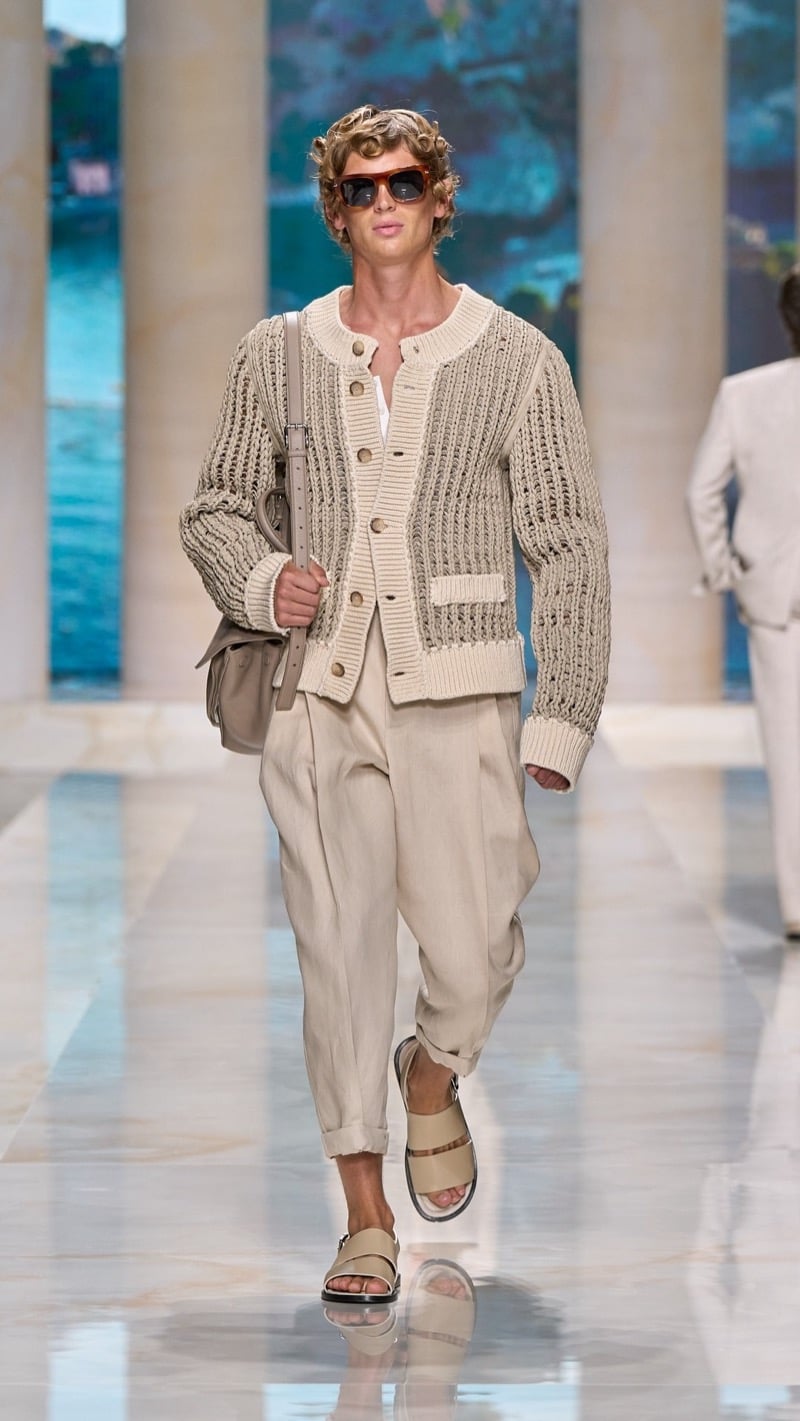 Model in tortoise sunglasses wears a chunky openwork rib-knit cardigan with raffia detail, paired with wide pleated oatmeal linen trousers, light sandals with leather straps, and a small tan suede bag