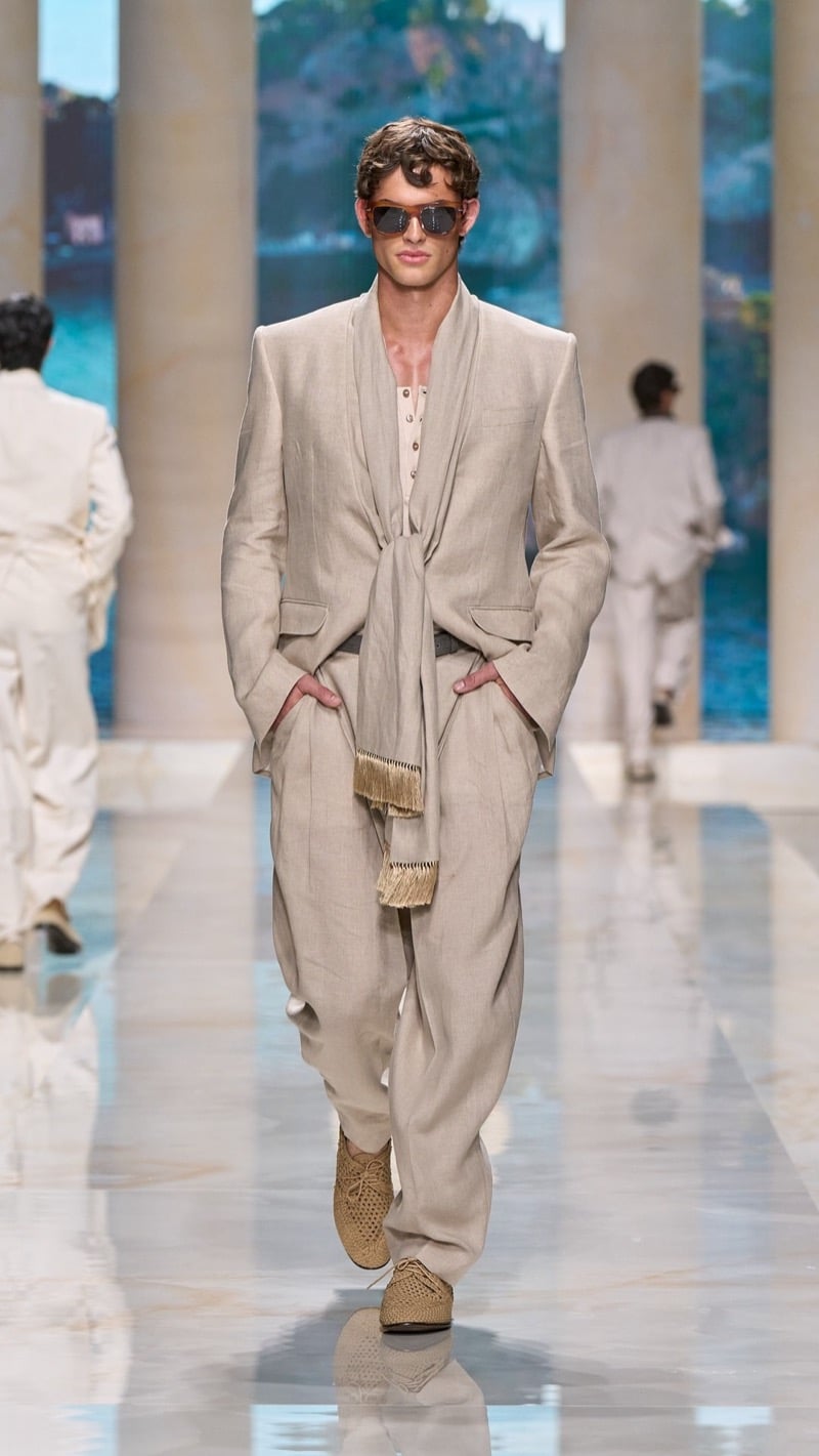 Model in tortoise sunglasses wears an oatmeal linen suit with a fringed scarf, layered over an oatmeal knit henley, paired with wide pleated trousers and raffia derbies