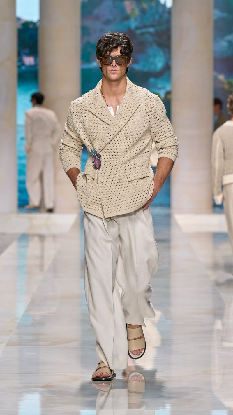 Model in shield sunglasses wears a double-breasted oversized openwork raffia knit jacket with a jeweled brooch, paired with wide pleated oatmeal linen trousers and beige slide sandals