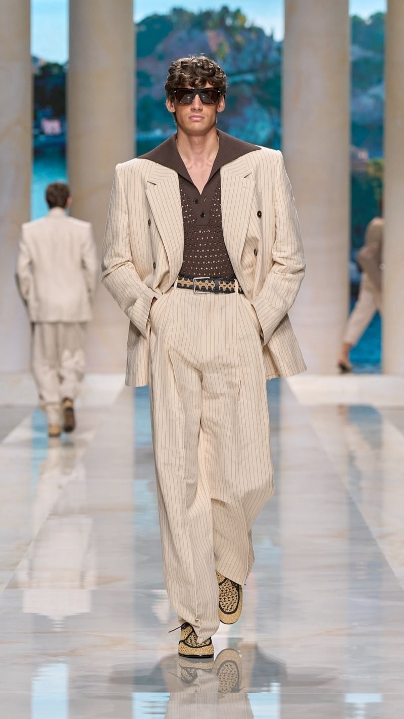 Model in shield sunglasses wears an oversized double-breasted oatmeal pinstripe suit with peaked lapels, layered over a chocolate brown crochet polo with rivet detail, paired with raffia derbies and an ornate buckled belt