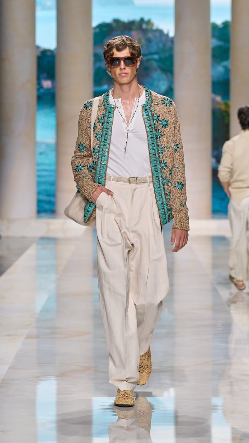 Model in tortoise sunglasses wears a raffia knit cardigan embellished with turquoise floral embroidery over a light henley with rosary necklace, paired with wide pleated oatmeal linen trousers, a woven belt, and raffia derbies